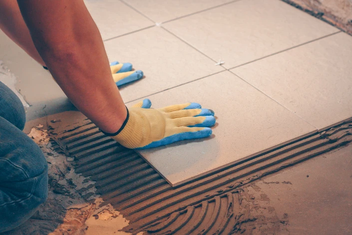 Travailleur en train de poser du carrelage sur sol intérieur, à genoux, portant des gants de protection jaunes et bleus